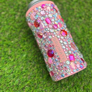 May include: A pink insulated can cooler bedazzled with a variety of colorful rhinestones. The cooler has a silver rim and the brand name "BRUMATE" is visible on the side. The rhinestones are in shades of pink, red, and clear.