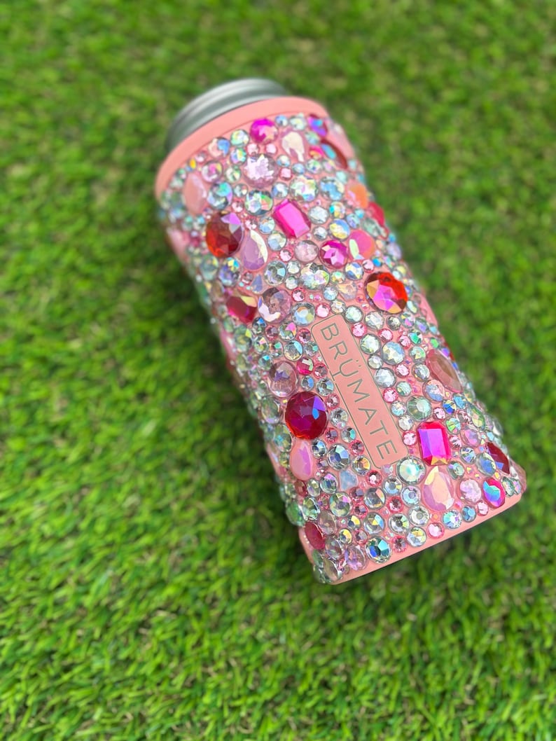 May include: A pink insulated can cooler bedazzled with a variety of colorful rhinestones. The cooler has a silver rim and the brand name "BRUMATE" is visible on the side. The rhinestones are in shades of pink, red, and clear.