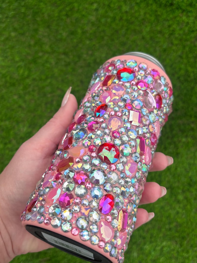 May include: A pink tumbler covered in a variety of colorful rhinestones. The rhinestones are in shades of pink, red, and iridescent colors. The tumbler has a smooth, glossy finish and a black base.