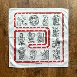 May include: White cotton bandana with a red border featuring a repeating pattern of footprints. The bandana has a central design of 12 black and white illustrations of figures and symbols from Mayan mythology. The text "TheJadeCircle.com" is printed in the bottom center of the bandana.