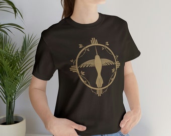 May include: A black t-shirt with a gold hummingbird design. The hummingbird is flying inside a circle with a stylized sun symbol and leaves.