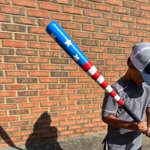 May include: A baseball bat with a patriotic design. The top of the bat is blue with white stars, transitioning to red and white stripes. The handle is black. The bat is held by a person in a gray shirt and a baseball cap.