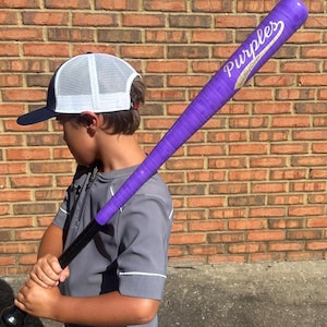 May include: A purple baseball bat with the word "Purples" printed on it. The bat has a black handle and a purple grip. The person in the image is wearing a gray shirt and a white and navy blue baseball cap.
