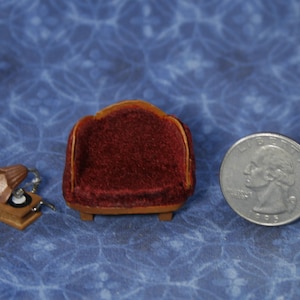 May include: Miniature dollhouse furniture set including a red velvet sofa, a small gramophone, and a quarter coin for scale. The sofa has a curved back and wooden base. The gramophone is brown with a silver horn.