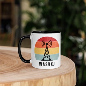 Puede incluir: Taza de cerámica blanca con asa e interior negros. La taza presenta un diseño retro de una torre de radio con ondas de señal, sobre un degradado de puesta de sol rojo, naranja, amarillo y azul. El texto "WA3VXJ" está impreso debajo.