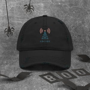 May include: Black distressed baseball cap with a teal and pink signal tower graphic and the word "ABCIDE". The cap has a curved brim and a distressed finish. The background has Halloween-themed decorations.