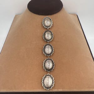 May include: A set of five oval-shaped silver-toned buttons arranged in a vertical column. Each button features a scalloped edge and a central diamond-shaped motif surrounded by radiating lines, creating a textured, metallic appearance throughout.