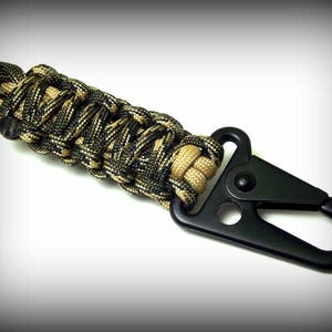 Puede incluir: Un llavero de paracord negro con un clip de metal negro. El paracord est&aacute; tejido en un patr&oacute;n de camuflaje con hilos negros y marrones.