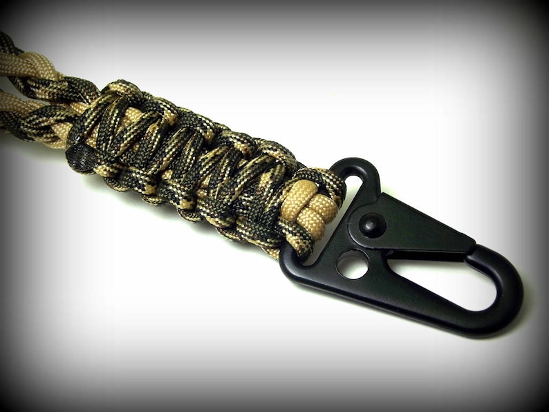 Puede incluir: Un llavero de paracord negro con un clip de metal negro. El paracord est&aacute; tejido en un patr&oacute;n de camuflaje con hilos negros y marrones.