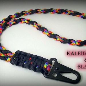 Puede incluir: Un cord&oacute;n de paracord negro y arco&iacute;ris con un clip de metal negro. El paracord est&aacute; trenzado con un patr&oacute;n negro y arco&iacute;ris. El texto "KALEIDOSCOPE & BLACK" es visible en la imagen.