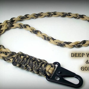 Puede incluir: Un cord&oacute;n de paracord de camuflaje negro y marr&oacute;n con un clip de metal negro. El cord&oacute;n tiene el texto "DEEP WOODS & GOLD" impreso en &eacute;l.