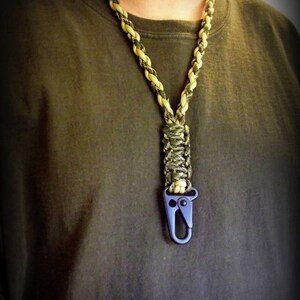 Puede incluir: Un cord&oacute;n de paracord negro y marr&oacute;n con un clip de metal negro. El cord&oacute;n se lleva alrededor del cuello de una persona.