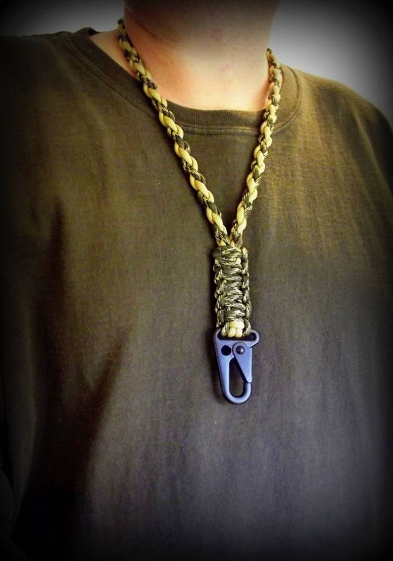 Puede incluir: Un cord&oacute;n de paracord negro y marr&oacute;n con un clip de metal negro. El cord&oacute;n se lleva alrededor del cuello de una persona.