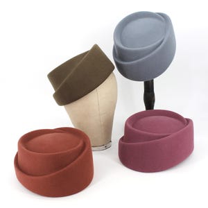 Peut inclure: Quatre chapeaux pillbox en vert olive, bleu ardoise, orange terre cuite et magenta profond. Le chapeau vert olive est présenté sur une tête de mannequin beige, tandis que les autres sont placés à proximité, dont un sur un support noir.