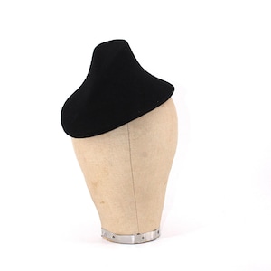Può includere: Un cappello nero dal design curvo unico, appoggiato su una testa di manichino. La forma del cappello ricorda un basco, con una corona leggermente rialzata. La testa del manichino è di colore beige neutro, in contrasto con il cappello.