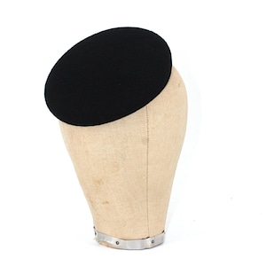 May include: A black, oval hat, possibly a pillbox or fascinator, is displayed on a beige mannequin head. The hat is made of a dark, solid material. The mannequin head is a neutral colour. The background is plain white.