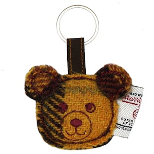 May include: A brown, yellow, and black tweed bear keychain with a brown leather strap and a silver key ring. The keychain has the text "Harris Tweed" and "Outer of Scotland 100% Pure New Wool" on a white tag.