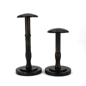 May include: Two dark brown wooden hat stands. Each stand has a round base, a central stem, and a rounded top. The taller stand has a decorative element on the stem. The stands are designed to display hats.