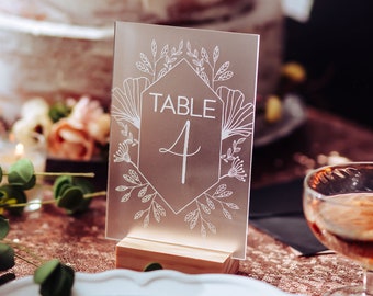 May include: A frosted acrylic table number with a floral design and the text "TABLE 4" in a white font. The number is on a wooden stand.