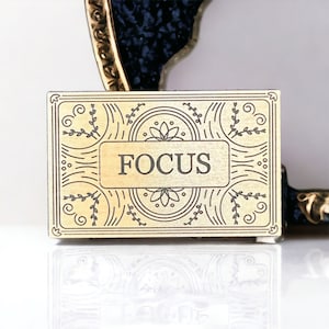 May include: A gold-colored metal plaque with an ornate design and the word "FOCUS" in black lettering.