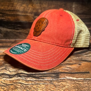 May include: A red and beige baseball cap with a brown leather patch featuring a skull with a forest design. The cap has a mesh back and the words "Wild & Free" and "Legacy" are printed on the front.