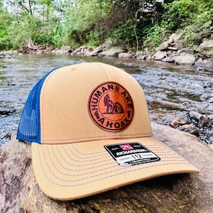 May include: A tan and blue trucker hat with a brown leather patch that says "Humans Are A Hoax". The hat is sitting on a rock near a stream.