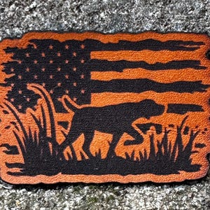 Op de afbeelding: Een rechthoekige patch met een Amerikaans vlagontwerp en een silhouet van een hond in hoog gras. De vlag heeft oranje strepen en een zwart sterrenveld. De hond en het gras zijn zwart en oranje.