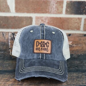 May include: A black and white distressed trucker hat with a brown leather patch that says "Dog Mom" with a paw print.