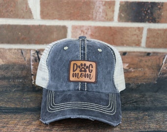May include: A black and white distressed trucker hat with a brown leather patch that says "Dog Mom" with a paw print.