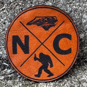 Może przedstawiać: Okrągła, pomarańczowa skórzana naszywka z czarną grafiką. Naszywka zawiera litery "NC", sylwetkę Karoliny Północnej i postać Sasquatcha. Czarne linie przecinają środek.