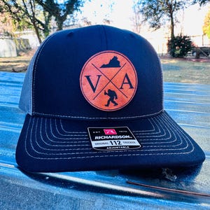 Könnte beinhalten: Eine schwarze und graue Trucker-Kappe mit einem braunen Leder-Patch, auf dem die Buchstaben "VA" und eine Silhouette eines Bigfoots zu sehen sind. Der Patch enthält auch eine Silhouette des Bundesstaates Virginia. Die Kappe hat einen weißen verstellbaren Riemen mit dem aufgedruckten Text "Richardson 112 Original Trucker Adjustable".