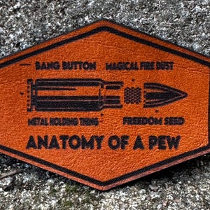 May include: An orange leather patch in a hexagonal shape, with black edging and text. The patch displays a bullet illustration and the phrases "BANG BUTTON," "MAGICAL FIRE DUST," "METAL HOLDING THING," "FREEDOM SEED," and "ANATOMY OF A PEW."
