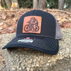 May include: Black and gray trucker hat with a brown leather patch featuring a cartoon monkey riding a mountain bike. The patch says "MTB".