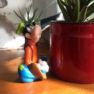 Op de afbeelding: Een keramiek beeldje van Goofy, een cartoonfiguur van Disney, zittend met gekruiste benen. Hij draagt ​​een groene hoed, een oranje shirt, een blauwe broek en witte handschoenen. Het beeldje staat op een houten tafel naast een rode pot met een groene vetplant.