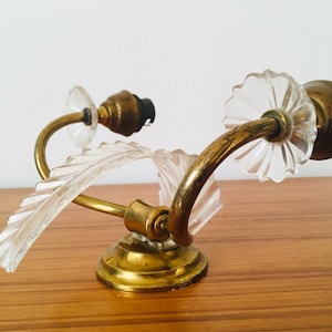 Może przedstawiać: Kinkiet ścienny z mosiądzu i szkła z dwoma oprawami. Kinkiet ma dekoracyjny wzór z przezroczystymi szklanymi liśćmi i mosiężną podstawą.