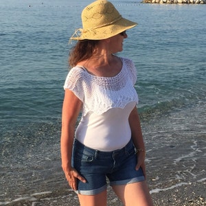 May include: A woman wearing a white crochet top, blue denim shorts, and a straw hat with a wide brim. She is standing on a beach with the ocean in the background.