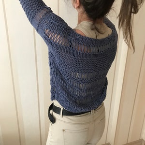 May include: A blue knitted sweater with a loose, open weave and a distressed look. The sweater has a scoop neck and long sleeves. The sweater is worn over a white tank top and paired with beige jeans.