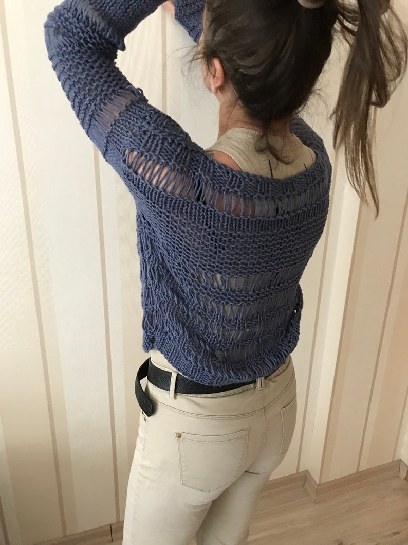 May include: A blue knitted sweater with a loose, open weave and a distressed look. The sweater has a scoop neck and long sleeves. The sweater is worn over a white tank top and paired with beige jeans.