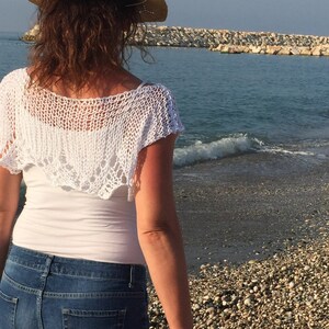 May include: A white crocheted shawl with a lacy design. The shawl is worn over a white tank top and blue jeans.