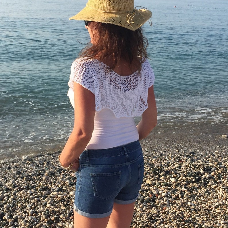 May include: A woman wearing a white crochet top, blue denim shorts, and a straw hat with a brown band. She is standing on a beach with the ocean in the background.