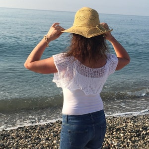 May include: A woman wearing a white crocheted top, blue jeans, and a straw hat with a wide brim. She is standing on a beach with the ocean in the background.