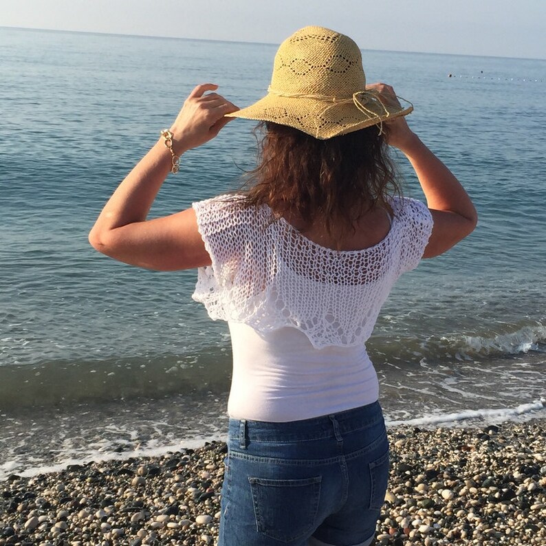 May include: A woman wearing a white crocheted top, blue jeans, and a straw hat with a wide brim. She is standing on a beach with the ocean in the background.