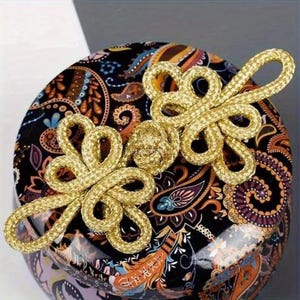 May include: Two gold-colored decorative knots rest on a round, patterned surface. The surface features a black background with a paisley design in shades of orange, purple, and blue. The knots are intricately woven.