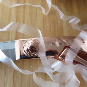 May include: Close-up of rose gold scissors with a decorative handle and silver blades. Sheer white ribbon is draped across the scissors and a light brown wooden surface.