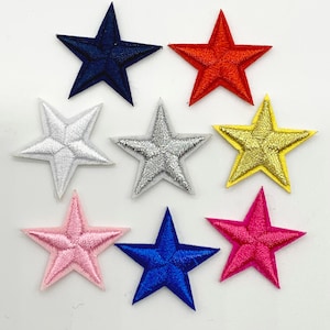 Może przedstawiać: Osiem haftowanych gwiazd z filcu w różnych kolorach, w tym czerwonym, niebieskim, złotym, srebrnym, różowym i białym. Gwiazdy są wszystkie tego samego rozmiaru i kształtu i są ułożone w losowym wzorze.