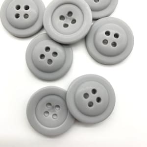 Könnte beinhalten: Eine Gruppe von sechs grauen Plastikknöpfen mit jeweils vier Löchern. Die Knöpfe liegen auf einer weißen Oberfläche.