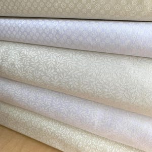 May include: A stack of five folded fabric bolts featuring light neutral tones. The patterns include repeating geometric pentagons, delicate leafy botanical prints, and small floral motifs in shades of cream, off-white, and pale beige.
