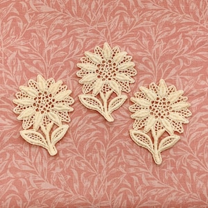 Könnte beinhalten: Drei cremefarbene Sonnenblumen-Applikationen aus Spitze. Jede Applikation weist detaillierte Blütenblatt- und Blattmuster auf. Die Applikationen sind auf einem rosa Stoff mit Blumenmuster angeordnet und erzeugen eine zarte und Vintage-Ästhetik.