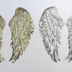 May include: Two pairs of sequined angel wings. One pair is gold, the other is silver. The wings are detailed with individual feather shapes and are likely for costume or craft projects. The wings are displayed on a white background.