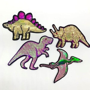 May include: Four sequined dinosaur patches in gold, purple, and green. The patches include a Stegosaurus, a Tyrannosaurus Rex, a Triceratops, and a Pterodactyl. Each patch has a black border.
